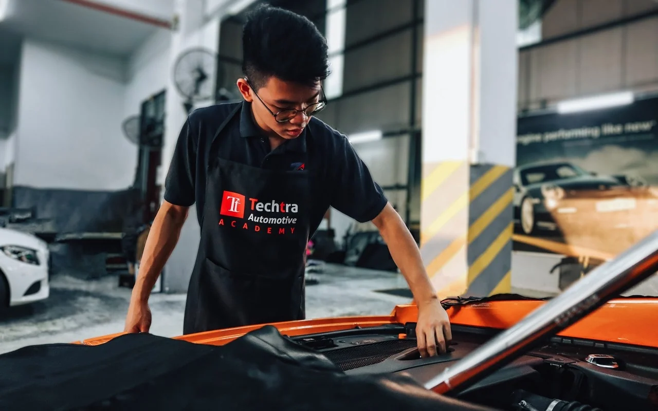 Seorang individu memakai apron Techtra Automotive Academy memeriksa kenderaan berwarna jingga di bengkel
